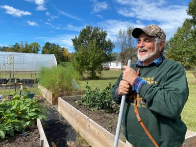 Blind farmer standing in front of raised garden beds. He is holding the handle of a garden tool and smiling.